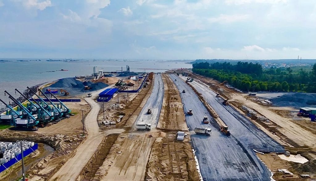 Hình ảnh thực tế công trường dự án với các tuyến đường san lấp, xe ủi và xe lu hoạt động liên tục, vật liệu xây dựng tập kết dọc bờ biển để thi công hạ tầng nền móng.